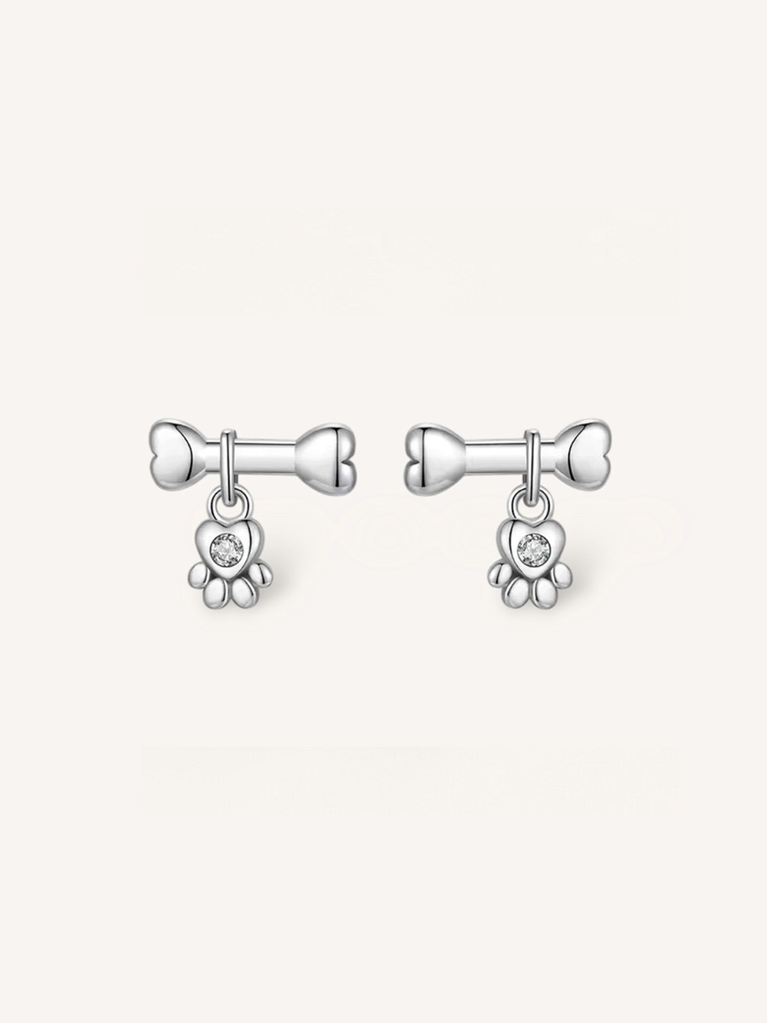 Pendientes pequeños de plata de ley con diseño de hueso del que cuelga una huella en forma de corazon con circonita central.