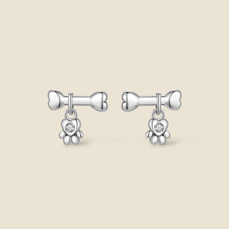 Pendientes pequeños de plata de ley con diseño de hueso del que cuelga una huella en forma de corazon con circonita central.